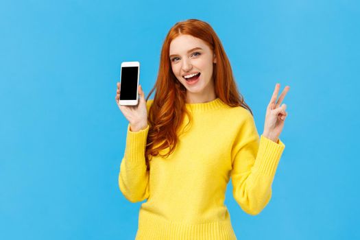 Positive and excited, charming redhead woman in yellow sweater, showing smartphone screen, advertise online store, mobile application, make peace sign and smiling joyfully, blue background.