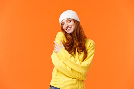 Tenderness, winter holidays concept. Charming romantic redhead woman in soft sweater, white beanie, close eyes and embrace own body, hugging herself, smiling cheerful, orange background.