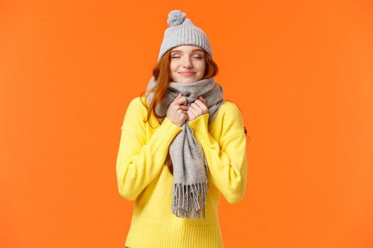 Cute and happy redhead woman walking along winter holidays fair market, close eyes and smiling as smelling something delicious, wearing warm grey hat and scarf, standing orange background.