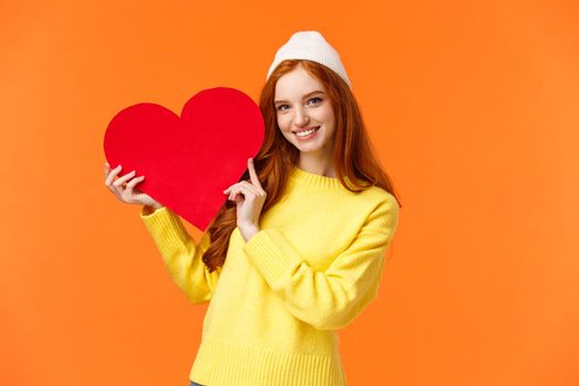 Relationship, boyfriend girlfriend and love concept. Cheerful lovely redhead girl holding big red heart sign and smiling, express affection, showing true-feelings to loved partner, orange background.
