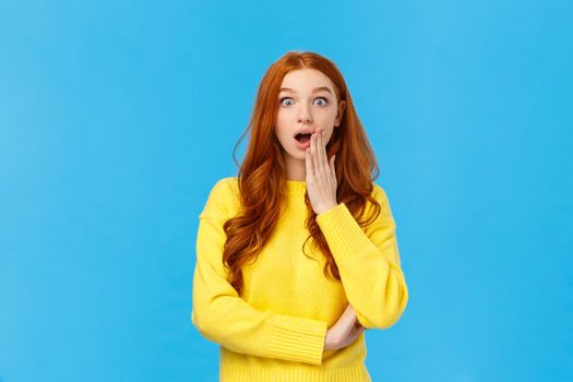 Wow stunning. Surprised girl found out secret or gossiping left shocked knowing truth. Astonished redhead woman stare camera speechless, gasping cover opened mouth amazed, blue background.