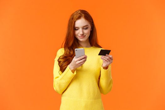 Waist-up portrait carefree modern young girl making online purchase, shopping internet using mobile application, insert credit card info smartphone, smiling, order something, orange background.