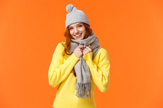 Its getting cold outside. Cute and lovely pretty redhead female wear grey winter hat and scarf, getting warm prepare play snowballs with friends outside during winter holidays, orange background.