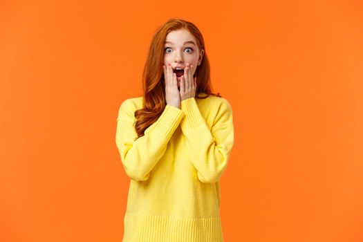 Impressed and speechless redhead woman found out awesome news, gasping, drop jaw astonished, touch cheeks and stare startled camera, christmas shopping season started, orange background.