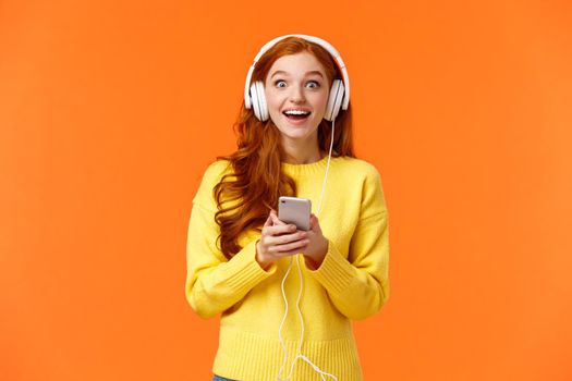 Surprised and astonished, excited smiling redhead girl found out favorite band released new song, listen music with fascinated, admiring eyes, grinning, holding smartphone, wear headphones.