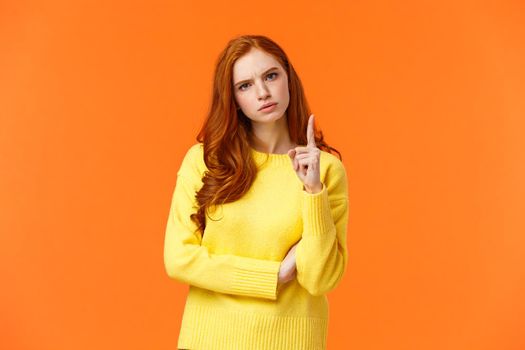 Redhead girl express disapproval as shaking her finger at someone with disappointed angry face, frowning give warning, prohibit unacceptable behaviour, standing displeased orange background.