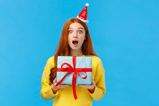 Surprised startled pretty redhead girl receive unexpected gift for christmas classmate, open mouth astonished and speechless staring camera as holding cute blue present wrapped box, studio background.