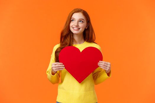 Dreamy cute redhead girl waiting for her prince, planning perfect velantines day romantic date, holding large heart sign and looking upper left corner with tender happy smile, orange background.