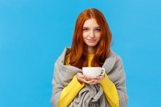 Redhead girl trying get warm, feeling cold wrapping herself with scarf on shoulders, drinking hot tea, holding teapot and smiling lovely, standing tender and cute blue background. Copy space