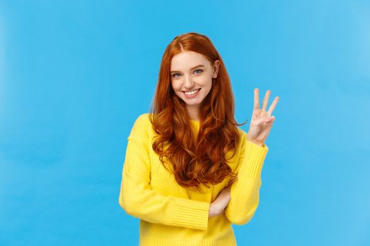 Gorgeous young redhead girl 20s in yellow sweater, smiling carefree making order, reservetion for three people, showing number third with fingers, standing blue background. Copy space