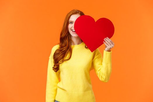 Holidays, romance concept. Cheerful girl showing affection, give heart to girlfriend, hiding half face behind valentines card, smiling joyfully, prepare romantic gesture for date, orange background.