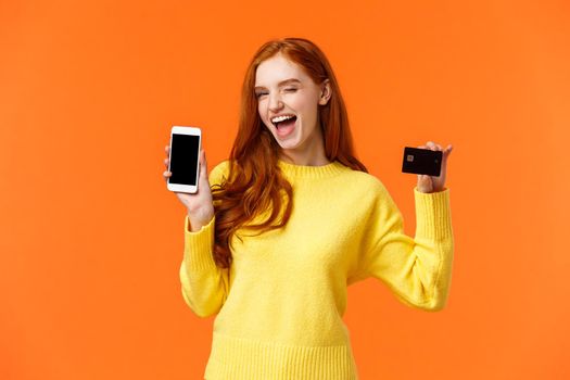 Shopping, advertising and mobile addication concept. Attractive cheeky redhead girl showing smartphone and credit card, wink and smile camera, promote online banking, deposit or payment method.