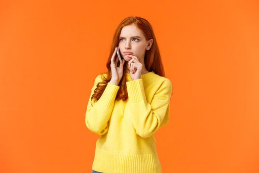 Indecisive and unsure, skeptical serious-looking redhead woman having tough decision make during conversation on phone, frowning touch lip pensive, order food delivery, orange background.