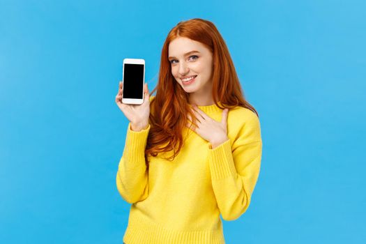 Impressed and fascinated redhead sassy woman touch heart and flirty gazing camera showing sexy guy photo she met dating app, holding smartphone with display facing camera, blue background.