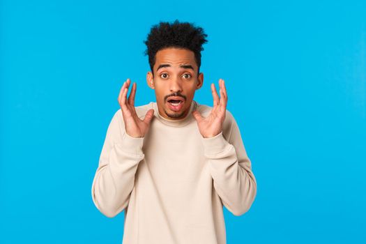 Man in panic telling something terrible, bad news. Alarmed, scared, shocked african-american guy witness accident, raising hands near face frightened and startled, gasping concerned, blue background.