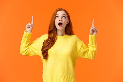 Wow amazing. Astonished and surprised redhead girl checking-out holiday special promo, woman stare and pointing up, stumble uppon cool offer, standing orange backgound astounded.