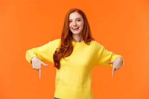 Waist-up portrait cheerful redhead female student in yellow sweater, inviting check-out event, recommend proudct, chrismas sale, holiday promo, pointing down and smiling, orange background.