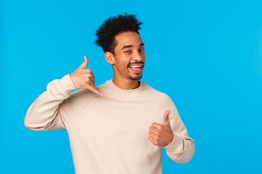 Call me later babe. Sassy and coquettish african-american confident macho guy with moustache, afro haircut showing thumb-up and phone gesture, wink and smiling flirty, blue background.