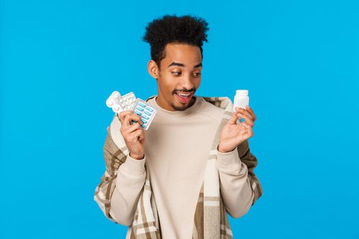 Funny handsome african man choosing what pill take first as got sick, staying home with cold or flu, holding pills from drugstore, use medication get better, taking care health, blue background.