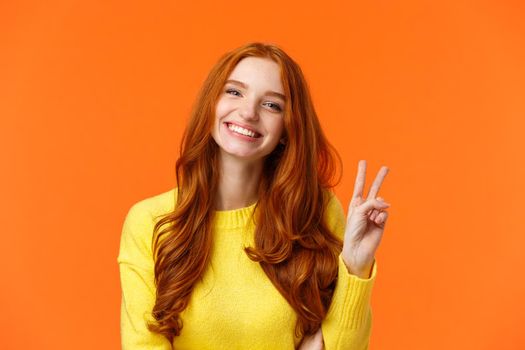 Waist-up cheerful gorgeous redhead woman with long curly red hair, showing peace sign and smiling happily, express positivity, like winter holidays, standing orange background.