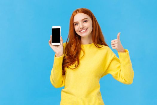 Girl showing good app, recommend download very useful application. Cute redhead woman in yellow sweater, like winter holiday sale in online store, showing smartphone display, blue background.