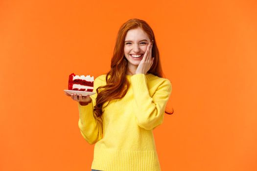 Happy lovely caucasian redhead female holding tasty cake with cream, touch cheek as receive congratulations being birthday girl, celebrating b-day with family, standing orange background.