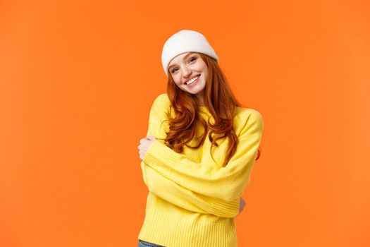 Winter is her favorite season. Tender silly and lovely redhead girl embrace herself and tilt head, smiling cute, wearing beanie and soft warm sweater just for cold weather, orange background.
