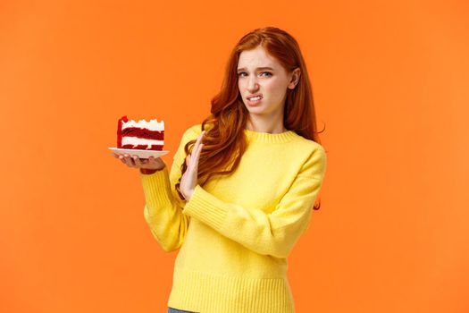 Disgusted and displeased, picky redhead girl look after body shape, dont eat sweets, holding piece tasty cake and refusing eat, showing stop sign, grimace at camera with aversion, orange background.
