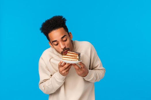 Tastes so good. Delighted and pleased african-american bearded guy likes sweets, attend friends birthday party, biting piece cake like taste, enjoy good food, standing blue background happy.