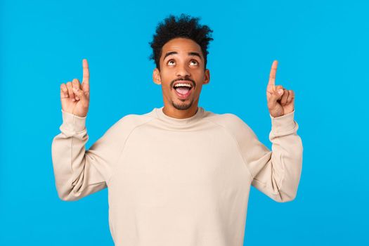 Guy seeing interesting sign, reating it from top. Attractive african-american male in winter sweater, pointing looking up and smiling impressed, happy found excellent promo, blue background.