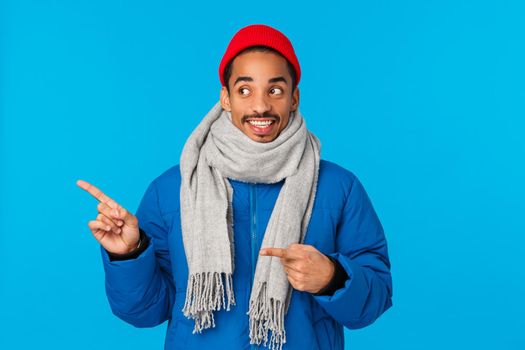 Wonder whats there. Cute and curious smiling happy african american guy in street wear, padded jacket, beanie and scarf, pointing glancing left with cheerful grin, standing blue background.