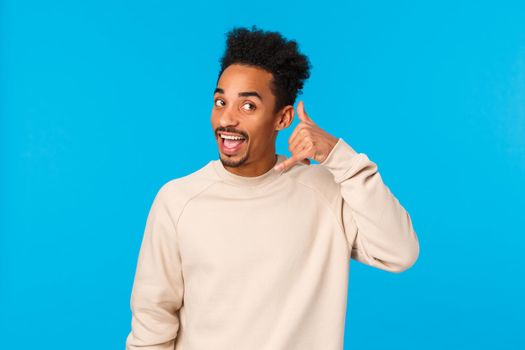 Hello whos calling. Funny attractive and cheerful hipster african american guy with afro haircut, moustache, imitating phone-call making mobile gesture near ear and answer, blue background.