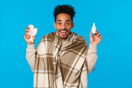 Cheerful guy feeling better after using prescribed medication doctor, wearing blanket as got sick, caught cold or flu, holding bottle nose spray and pills from drugstore, smiling, blue background.