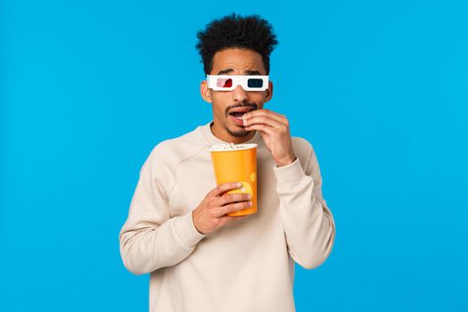 Scared and entertained, intrigued african american silly guy watching 3d movie in cinema, afraid of looking screen but interested what happen next, eating popcorn and stooping afraid, blue background.