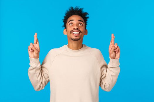 Enthusiastic and intrigued modern african-american male in white sweater, seeing something curious upwards, look and pointing fingers up, smiling astonished, wonder whats there, blue background.