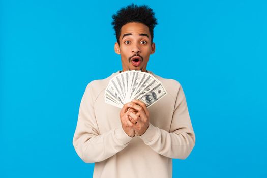 Lottery, sport bet and finance concept. Amazed and startled happy african-american guy got lots of cash, winning prize, holding big money and smiling, gasping astonished, blue background.