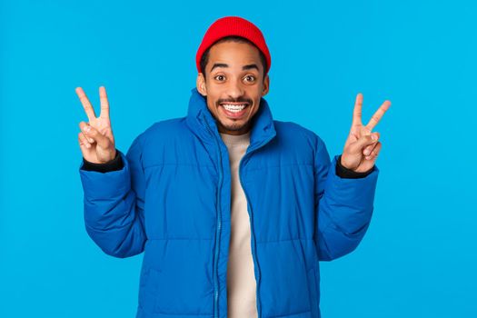 Say cheeze. Ecstatic cheerful young african american guy enjoy winter holidays, like christmas and new year, standing in padded jacket, red beanie, showing peace gestures and smiling, blue background.