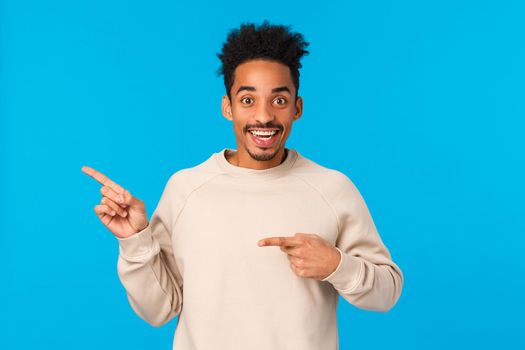 Surprised, excited african-american guy with afro haircut asking about product, interested in event pointing left and looking camera thrilled, cant wait celebrate new year party, blue background.