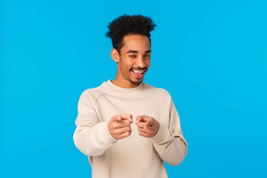 You rock, congratulations. Sassy good-looking african american hipster guy pointing fingers at camera and winking as choosing, indicating at winner or lucky person who achieved goal, blue background.