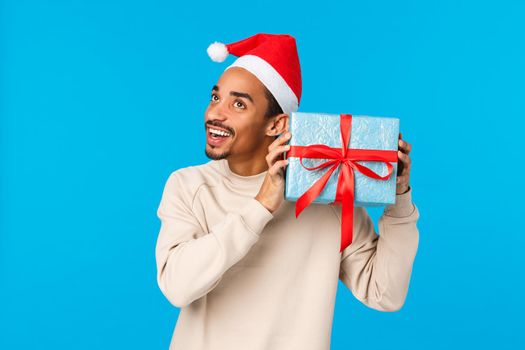 Hmm interesting whats inside. Curious and enthusiastic happy african-american guy in santa christmas hat, shaking gift box near ear to hear and guess what is it, smiling, celebrating winter holidays.