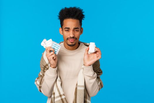 Health, medication and winter season concept. Young african american guy holding drugs and bottle of pills, sneezing, feel unwell fatigue from flu, caught cold, standing in blanket blue background.