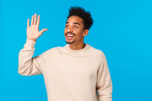 Hey hello, over here. Attractive friendly african-american hipster guy afro haircut, moustache, seeing friend in crowd, looking upper left corner, waving hand in hi gesture, smiling, blue background.