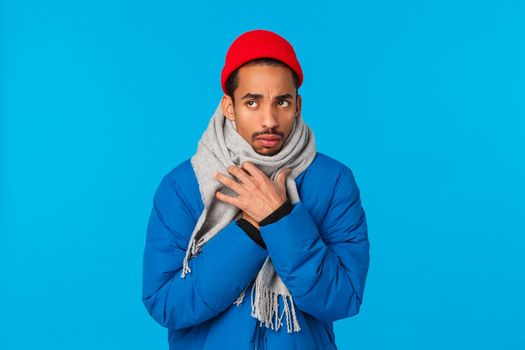Silly and sad, moody african-american guy acting childish sulking, looking upset upper left corner, feeling cold as standing windy snowing weather, wrap himself scarf, wear padded jacket, red beanie.