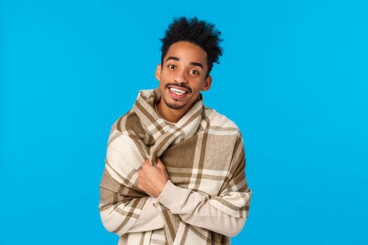 Man trying get warm asking for tea as staying home after walking on winter cold weather, wrap himself in blanket and smiling grateful, visit granny for christmas holidays, blue background.