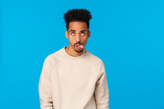 Waist-up shot moody and home-sick handsome african-american guy fooling around, feeling bored and unamused showing tongue, grimacing rolling eyes up, standing blue background careless.