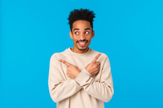 Guy making assumption what he get for holidays. Cute and excited charming african american hipster male in white sweater, pointing sideways left and right, smiling eager start celebrating, blue wall.