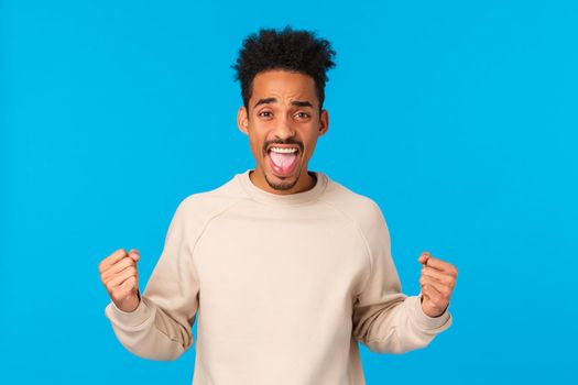Cheerful and excited handsome african-american male with afro haircut, clench fists showing strength power, triumphing, rooting for team, watching sport game and cheering, blue background.