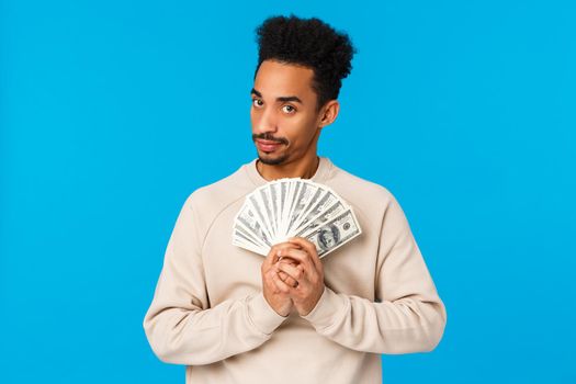 Guy got cash, thinking what buy, how waste money during winter holidays sale season. African american good-looking man with afro haircut holding dollars near chest and gazing suspicious camera.