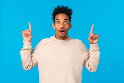Wow awesome. Impressed good-looking african guy with moustache and afro haircut, folding lips smiling and staring, pointing up at amazing breathtaking event, advertising, blue background.
