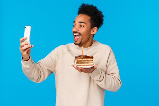 Excited happy african-american bearded hipster guy holding piece cake with birthday candle, smiling joyfully taking selfie or record video how he celebrates, making wish, standing blue background.